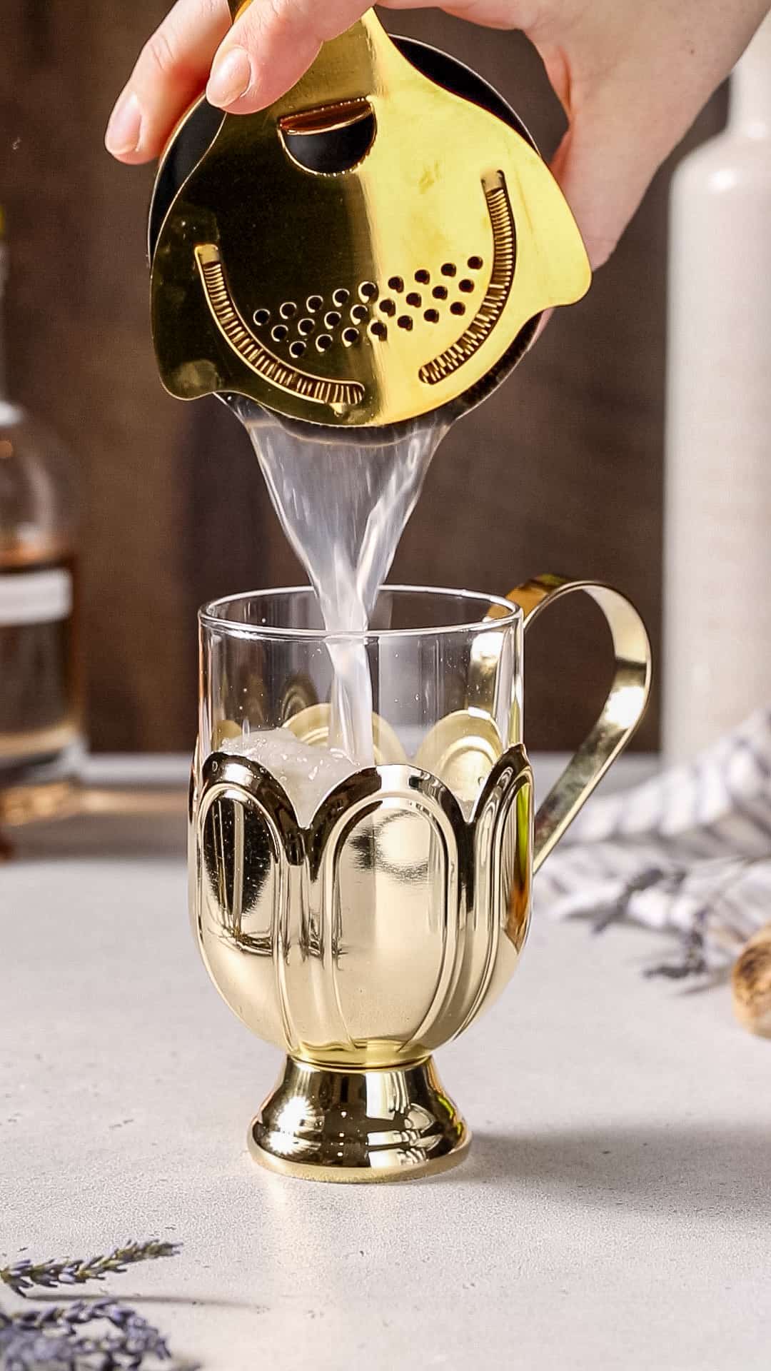 Hand pouring liquid into a gold and glass cocktail serving mug using a shaker and strainer.