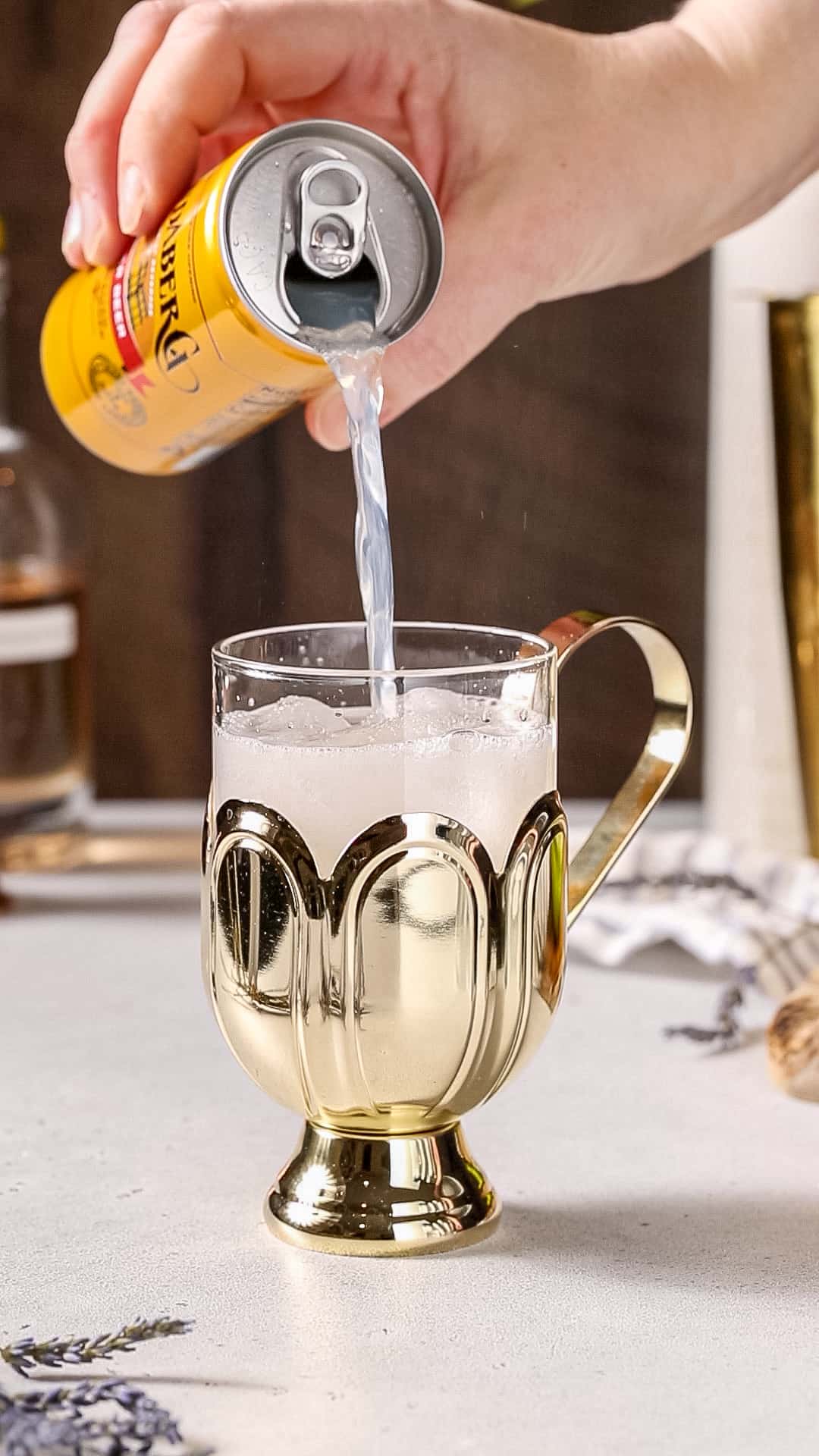 Hand pouring ginger beer into a cocktail mug filled with ice and some other liquid.