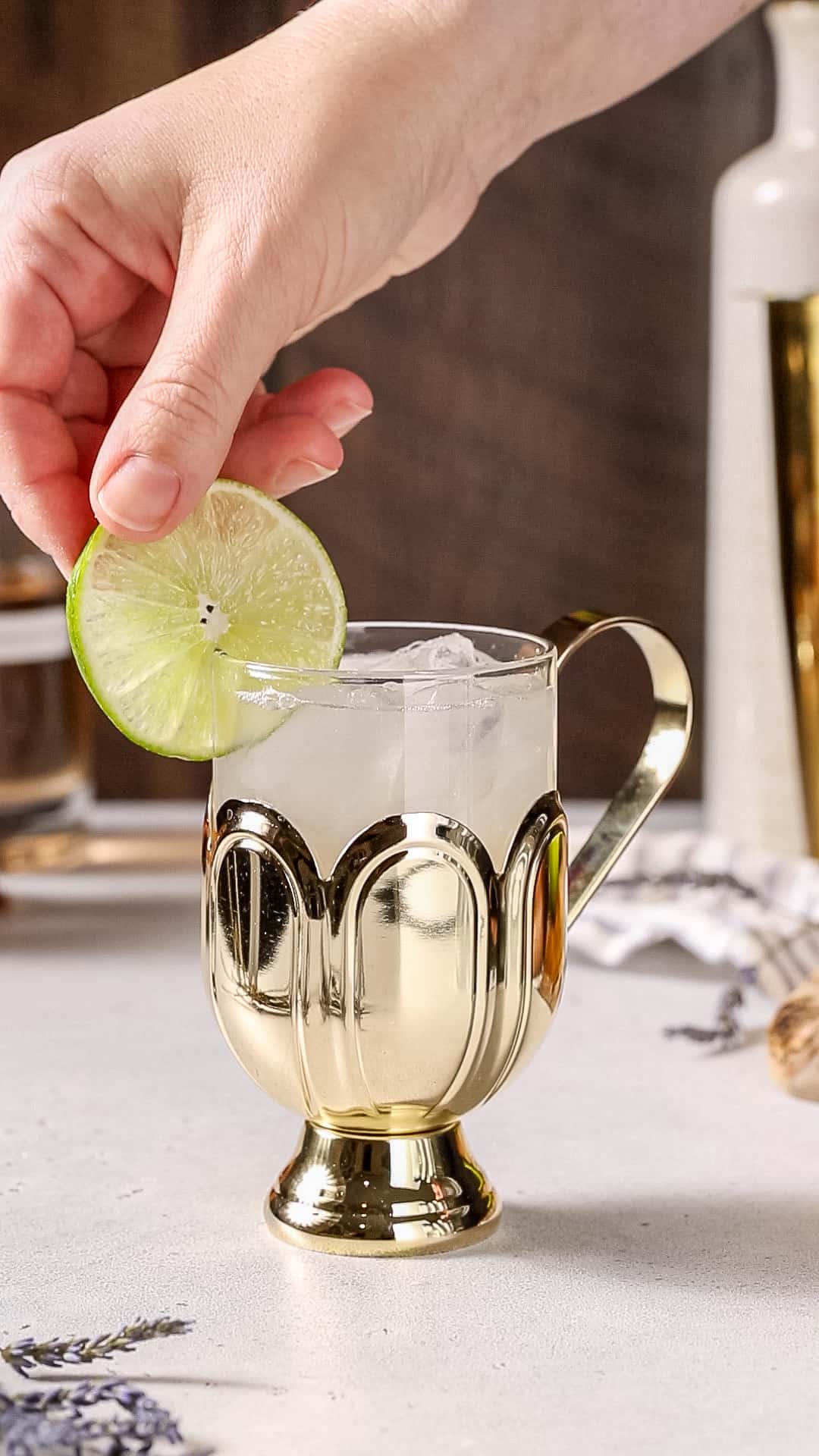 Hand adding a lime slice to the rim of a gold and glass cocktail mug.