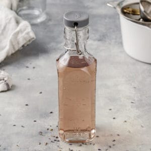 Lavender syrup in a sealed bottle on a gray countertop. There is a pot, funnel and linen in the background.