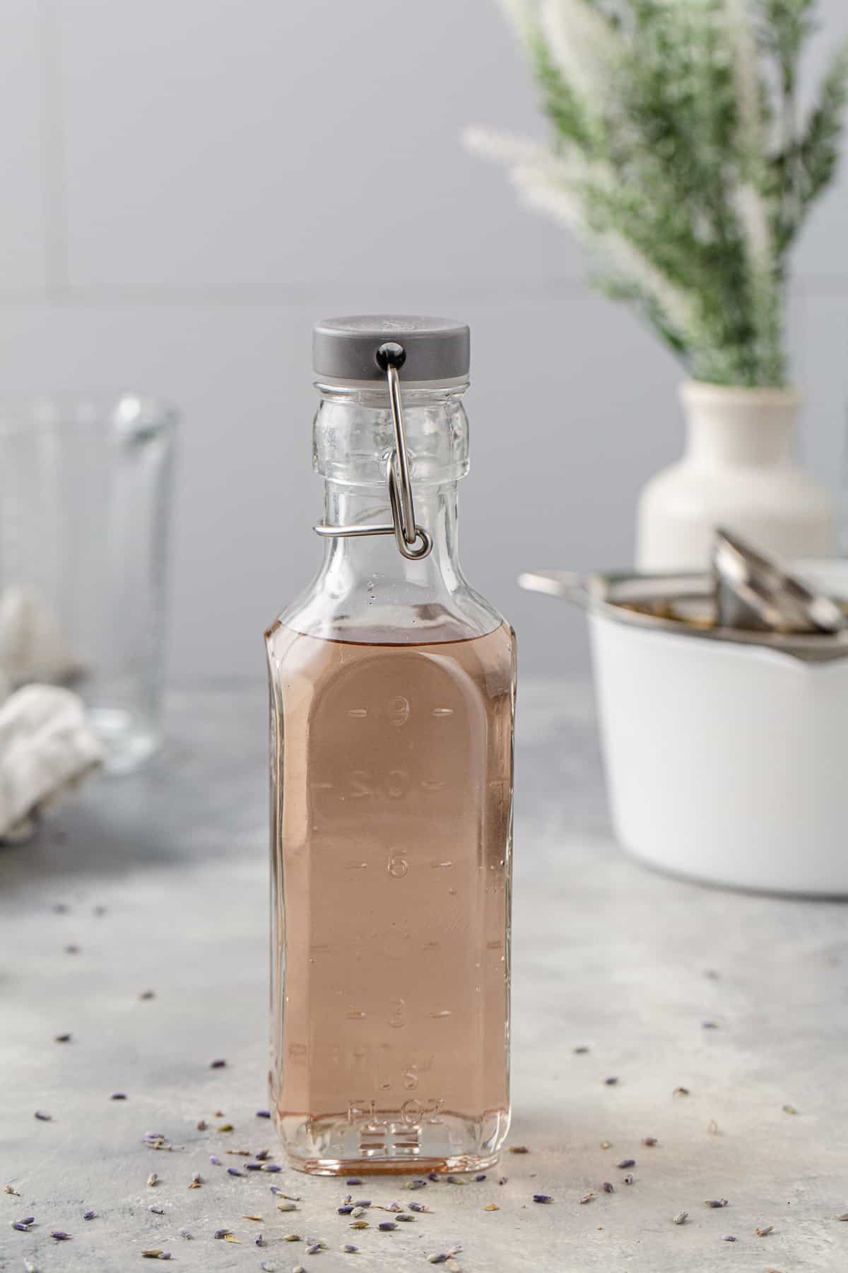 Bottle of lavender syrup on a gray countertop. In the background is a white pot and other tools to make the syrup, along with a vase of greenery.