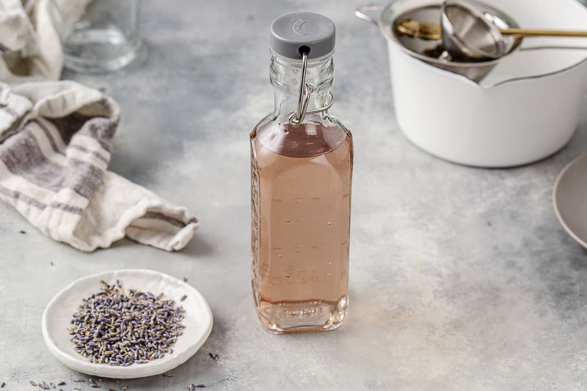 Overhead view of lavender syrup bottle on a gray countertop. Equipment to make the syrup is off to the side and there is a small dish of dried lavender buds to the left.