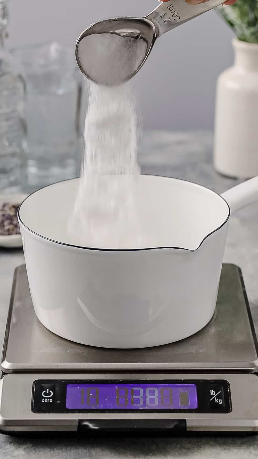 Hand pouring sugar from a measuring spoon into a white pot.