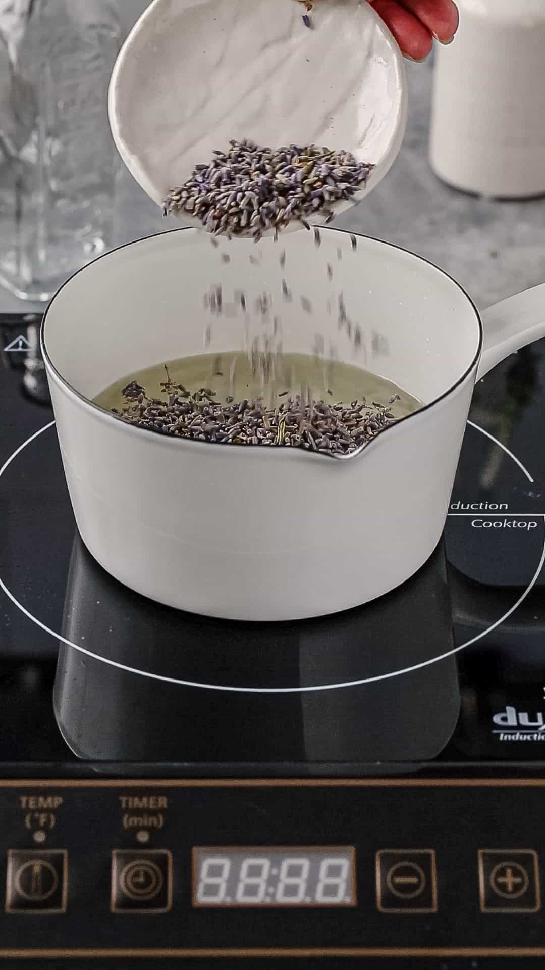 Hand adding dried lavender from a small dish into a white pot filled with hot syrup.