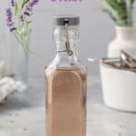 Bottle of lavender syrup on a gray countertop. In the background is a white pot and other tools to make the syrup, along with a vase of greenery. Text at the top reads “Lavender Syrup” in purple and a sprig of lavender illustration is next to the text.