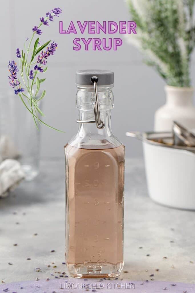 Bottle of lavender syrup on a gray countertop. In the background is a white pot and other tools to make the syrup, along with a vase of greenery. Text at the top reads “Lavender Syrup” in purple and a sprig of lavender illustration is next to the text.