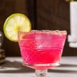 Pink colored prickly pear margarita in a salt rimmed glass garnished with a lime wheel. A cactus can be seen in the background on the right.