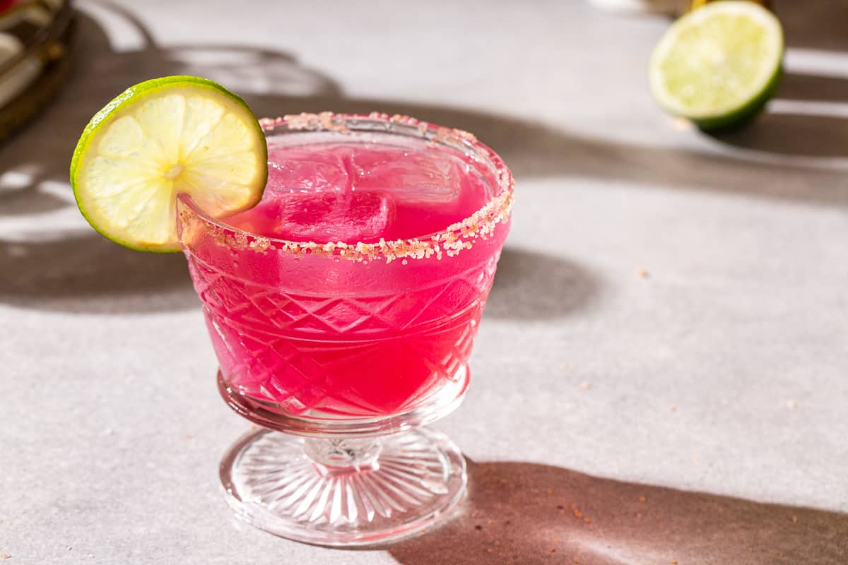 Pink colored prickly pear margarita in a salt rimmed glass garnished with a lime wheel. Half of a lime is in the background to the right.