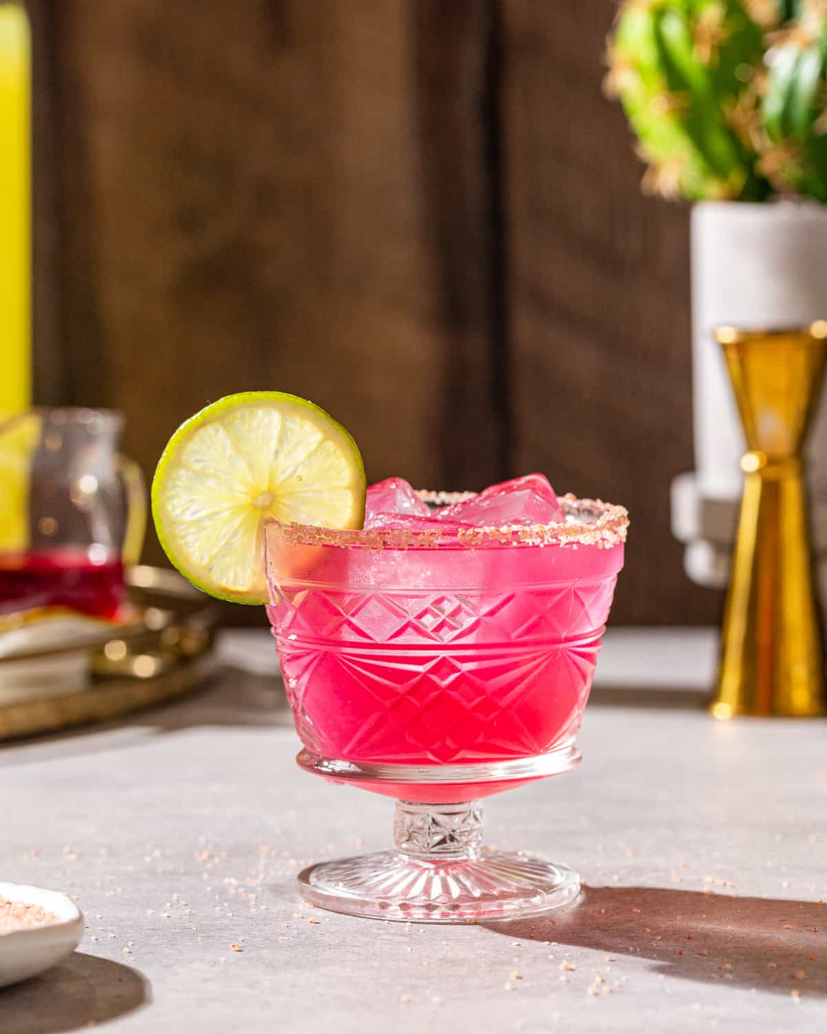 Side view of a pink colored prickly pear margarita in a short-stemmed serving glass with a salted rim. There is a jigger, cactus, bottle of Limoncello and a small jug of prickly pear syrup in the background.