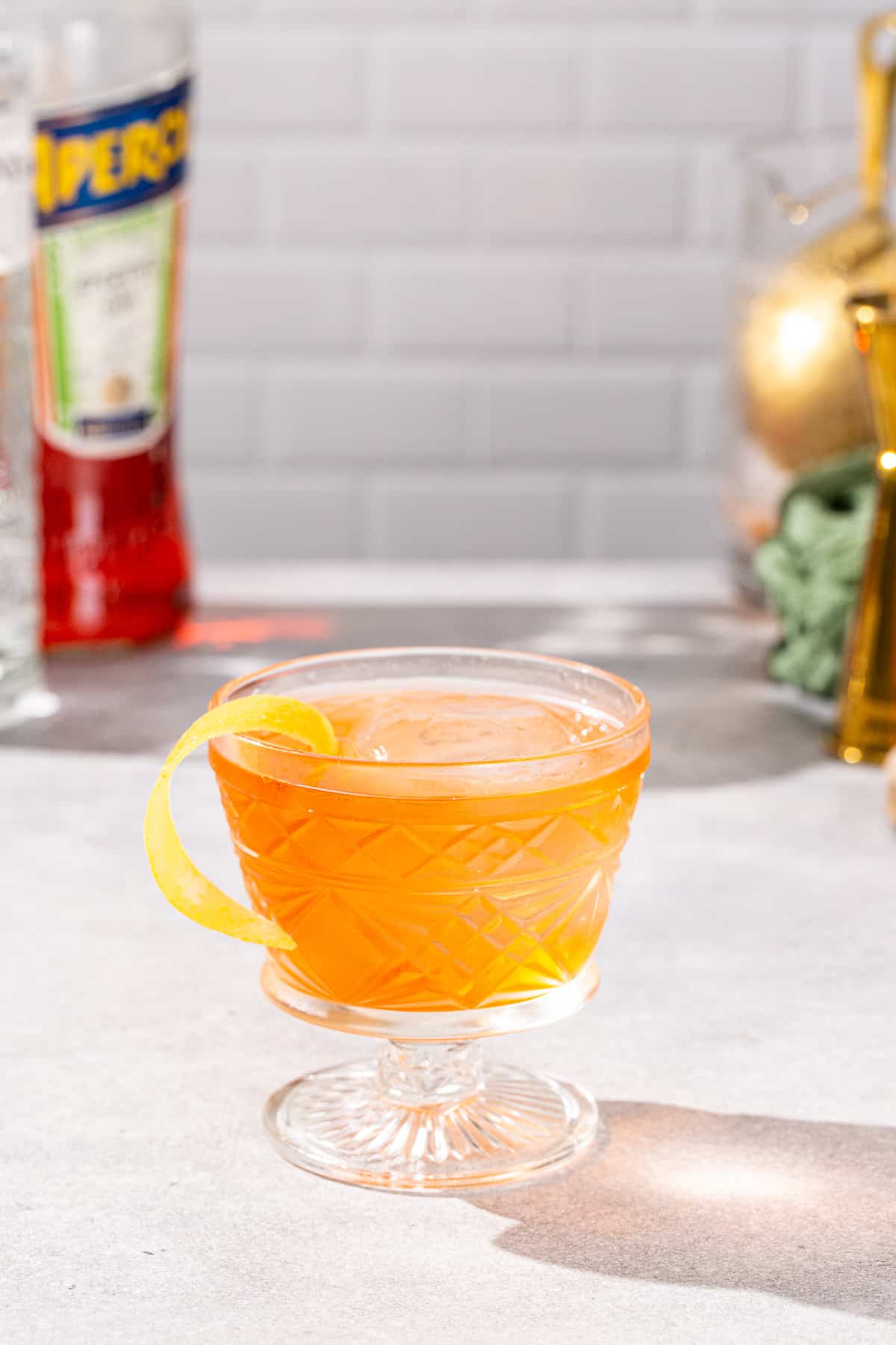 Side view of Aperol Negroni cocktail in a vintage style cocktail glass with a short stem. The drink is orange colored and has a yellow lemon peel garnish. A bottle of gin and aperol are visible in the background along with bar tools.