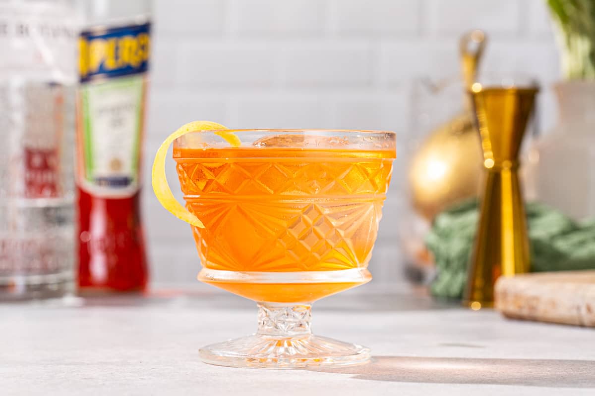 Side view of Aperol Negroni cocktail in a vintage style cocktail glass with a short stem. The drink is orange colored and has a yellow lemon peel garnish. A bottle of aperol is visible in the background along with bar tools.