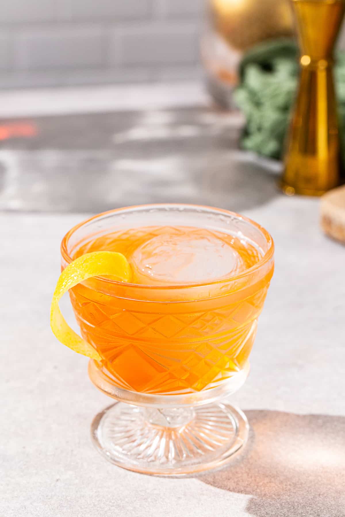 Slight overhead view of Aperol Negroni cocktail in a vintage style cocktail glass. The drink is orange colored and has a yellow lemon peel garnish. A gold colored jigger and a wood cutting board are visible in the background.
