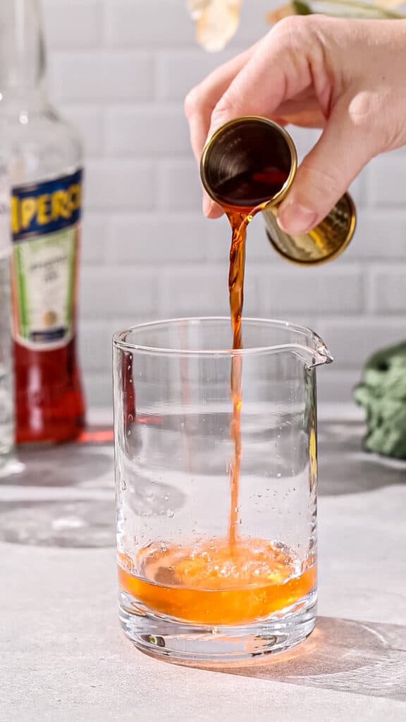 Hands using a jigger to pour aperol amaro liqueur into a cocktail mixing glass.