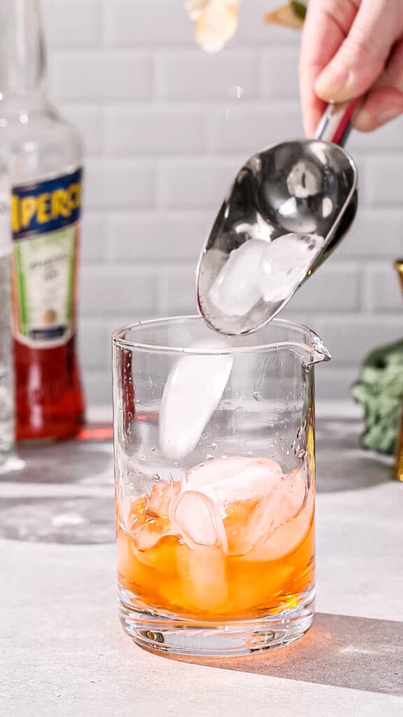 Hand using an ice scoop to add ice to a cocktail mixing glass filled with orange liquid.