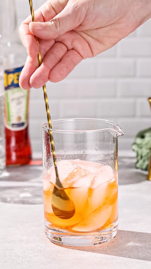 Hand using a bar spoon to stir down an orange cocktail with ice.