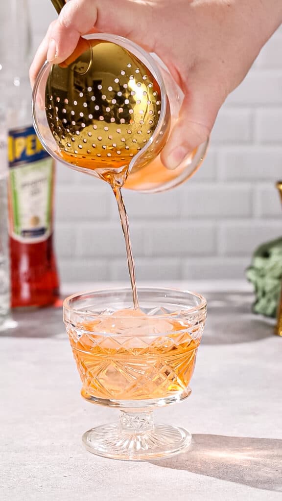 Hand straining an orange drink into a vintage cocktail serving glass.