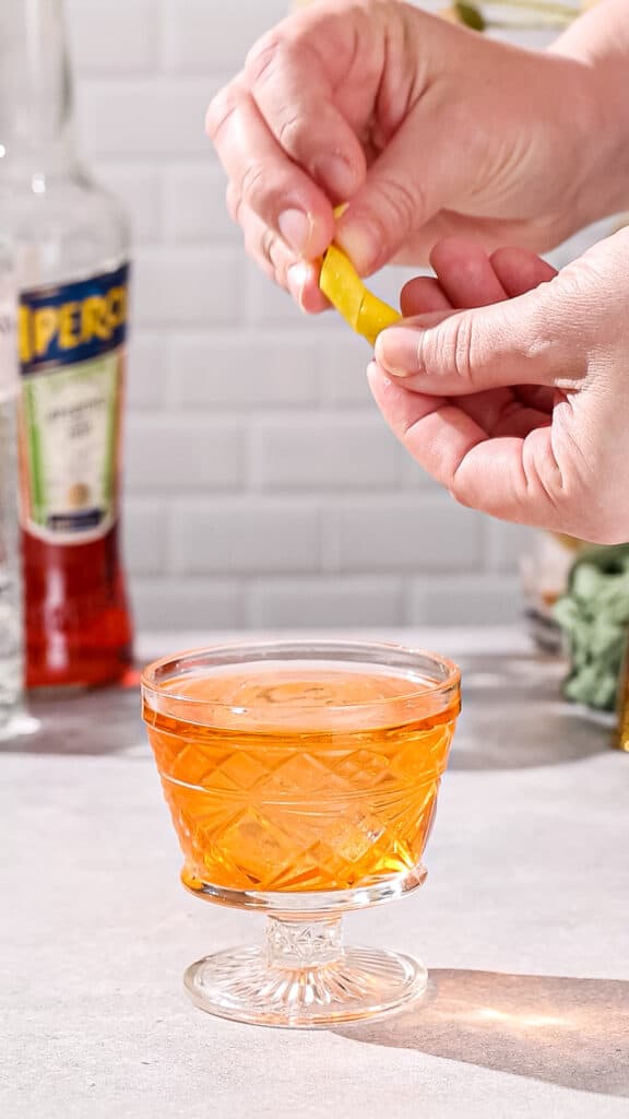 Hands twisting lemon peel over top of an orange cocktail.