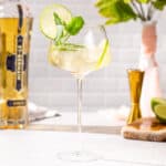 Hugo Spritz cocktail in a tall stemmed cocktail glass. A bottle of St Germain is in the background along with cut limes, a jigger and a vase. The drink is garnished with lime slices and fresh mint.