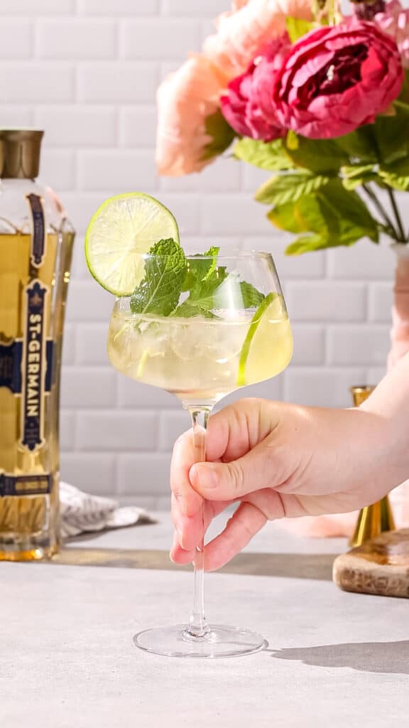 Hand about to pick up a Hugo spritz cocktail with fresh lime slices and fresh mint garnish.
