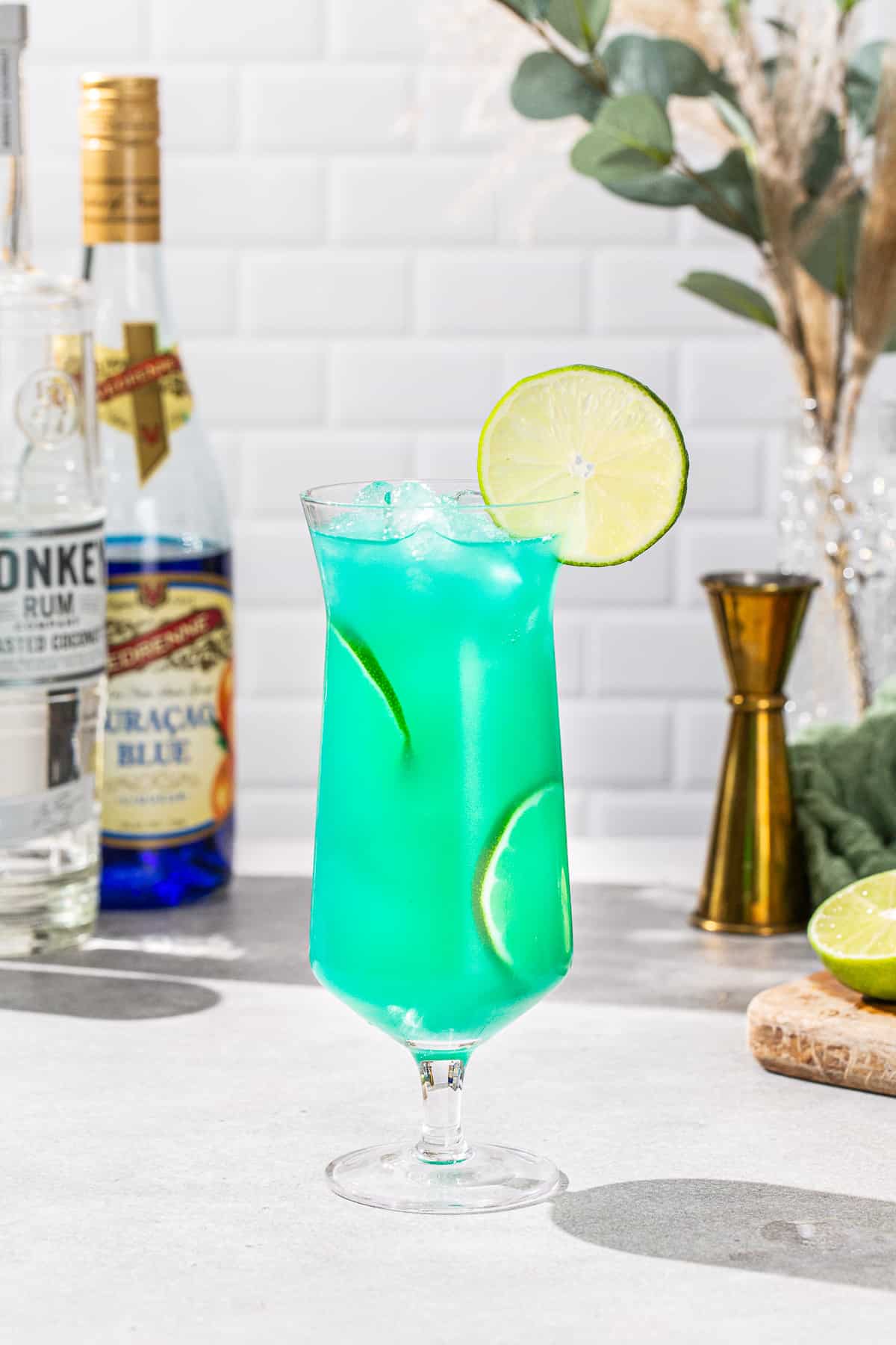 Side view of a Mermaid Water cocktail in a hurricane glass. The drink is vibrant aqua with lime slices as garnish. In the background are bar tools and ingredients to make the drink.