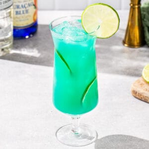 Slightly overhead view of a Mermaid Water cocktail in a hurricane glass. The drink is vibrant aqua with lime slices as garnish. In the background are a gold jigger as well as ingredients to make the drink.
