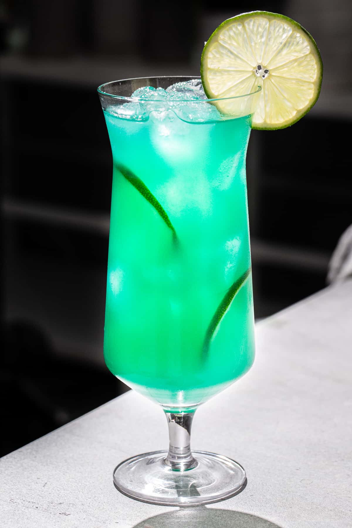 Side view of a Mermaid water cocktail with three cut lime slices as garnish. The drink is vibrant aqua in color. It is backlit on a darkened background with light coming from behind to light up the glass. The drink sits on a grey countertop.