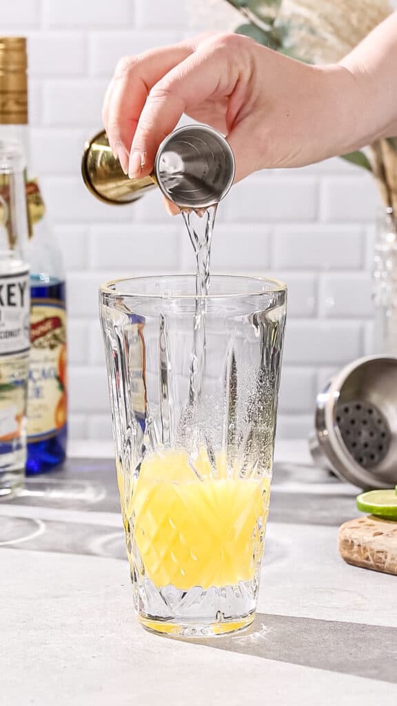 Hand adding vanilla vodka to a cocktail shaker filled with yellow liquid.