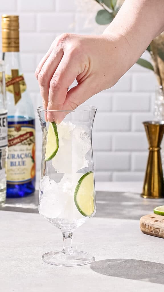 Hand adding lime slices to a hurricane glass filled with ice.