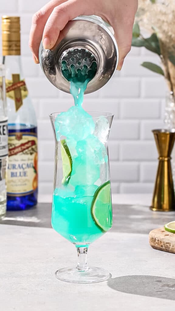 Blue liquid being poured from a cocktail shaker into a tall hurricane glass filled with ice and lime slices.