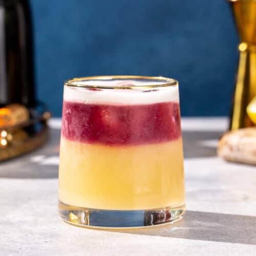 Side view of a New York Sour cocktail with a red wine float and egg white foam on top. In the background are a jigger and ingredients used to make the drink.