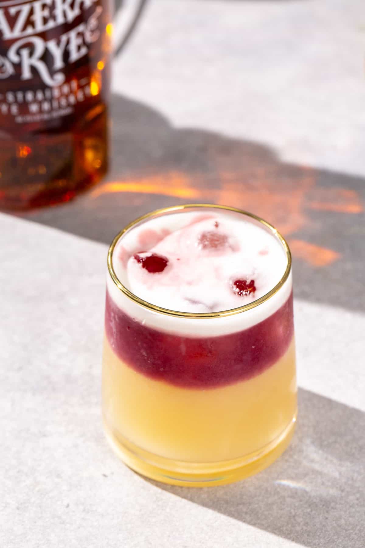 Overhead view of a New York Sour cocktail with a red wine float and egg white foam on top. A bottle of rye whiskey is in the background.