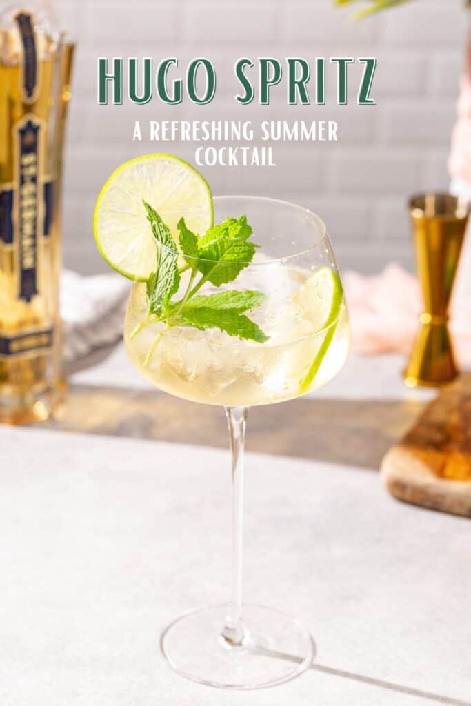 Slightly overhead view of Hugo Spritz cocktail in a tall stemmed spritz glass, with St-Germain liqueur in the background along with a jigger and cutting board. Text overlay says "Hugo Spritz, a refreshing summer cocktail".