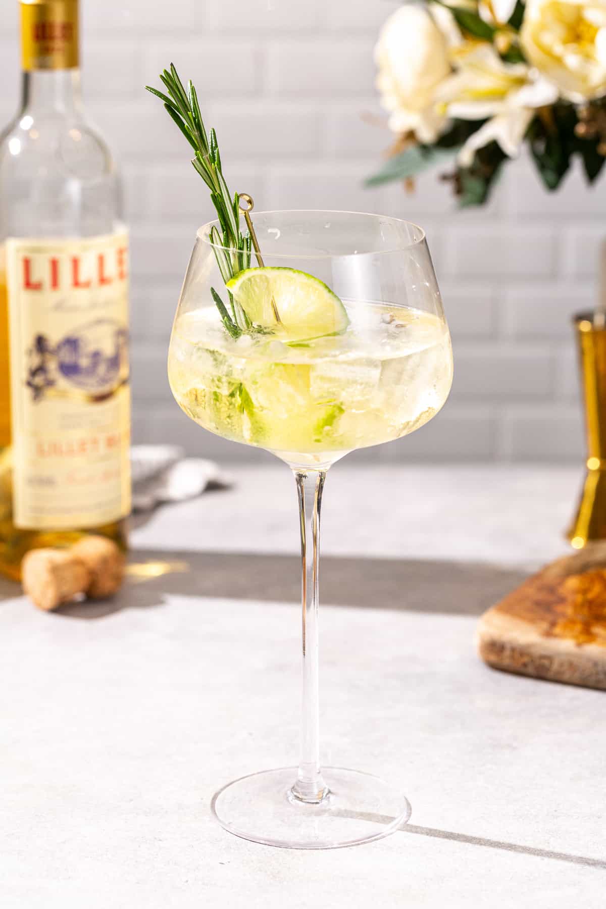 Side view of the LIllet Spritz cocktail in a tall stemmed glass. A lime slice and fresh rosemary garnish the drink. In the background are a bottle of Lillet Blanc, a prosecco cork and bar tools.