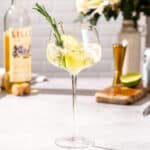 Side view of Lillet Spritz cocktail with a lime and rosemary garnish on a countertop. The drink looks bright and refreshing. Bar tools and bottle of Lillet are in the background along with a vase of flowers.