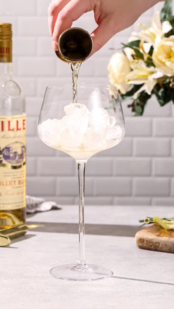 Hand pouring Lillet blanc from a jigger into a cocktail glass filled with ice.