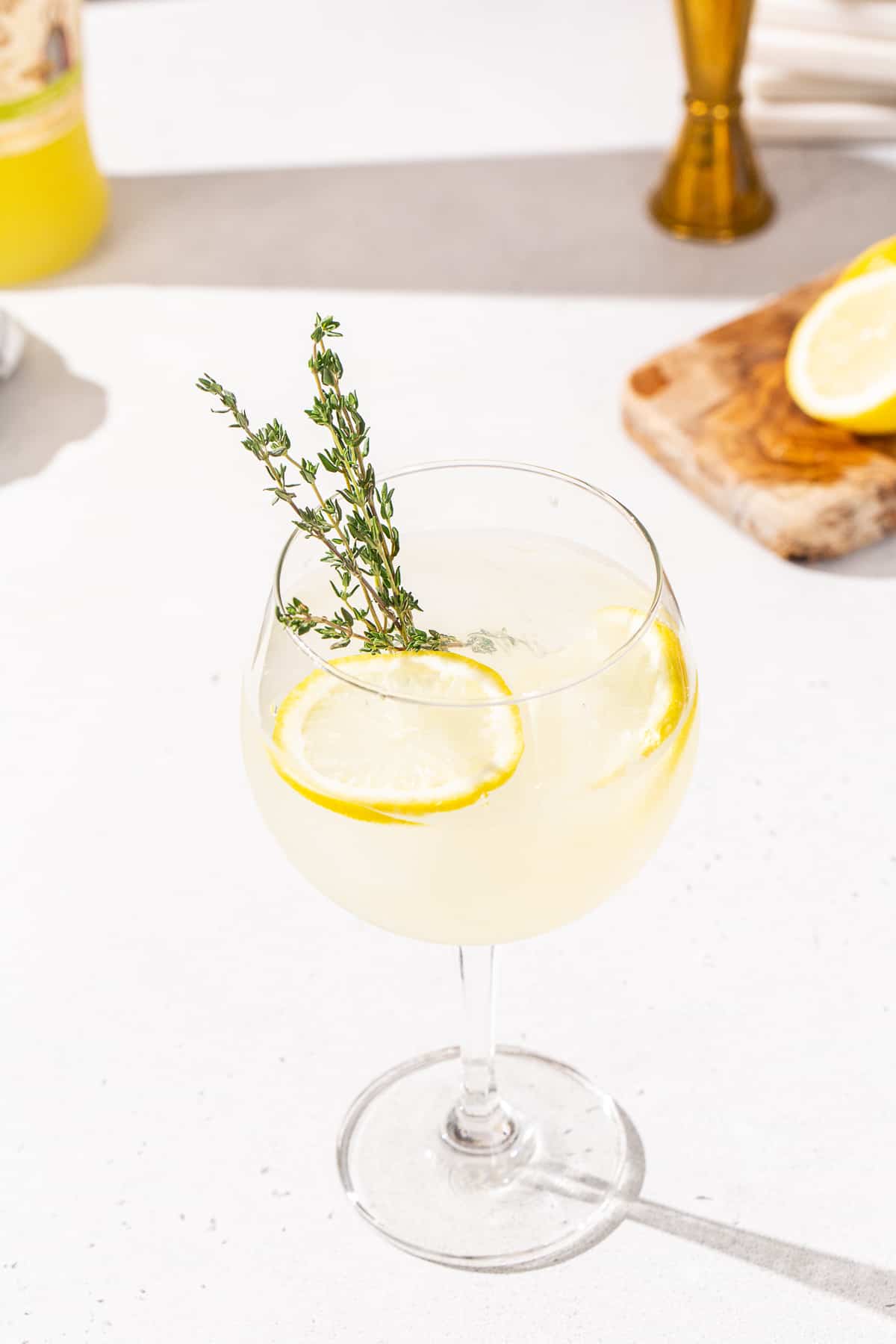 Slightly overhead view of Limoncello Spritz cocktail on a countertop. A bottle of limoncello, a jigger and a wood cutting board with cut lemon slices are visible in the background. The drink has a fresh thyme sprig and lemon slices as garnish.