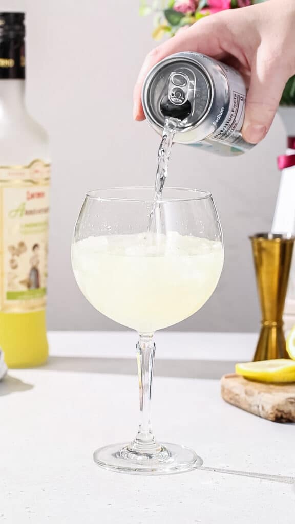Hand pouring seltzer into a cocktail glass filled with yellow liquid.