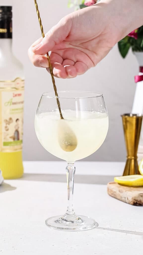 Hand using a gold colored bar spoon to gently stir limoncello cocktail.