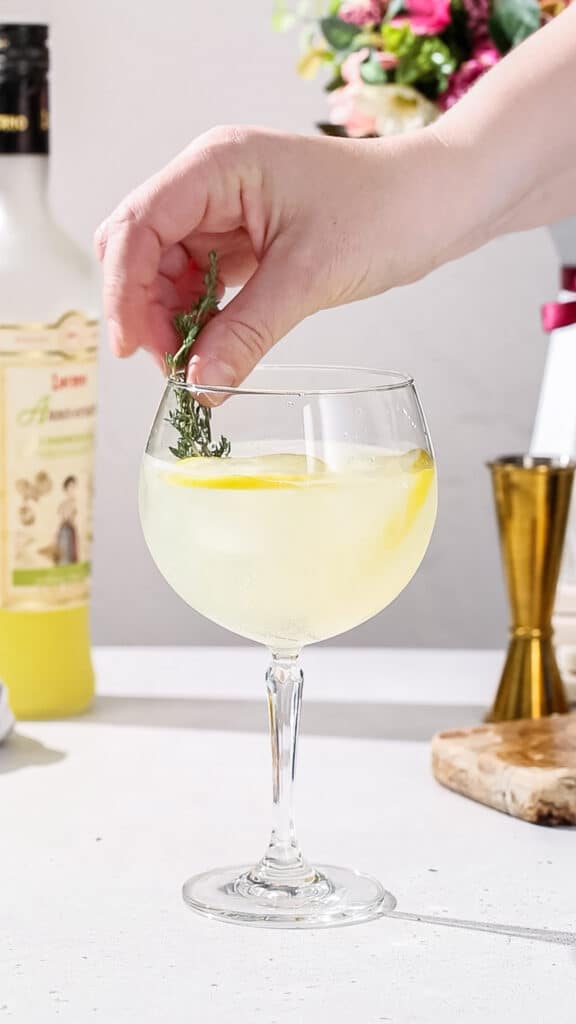 Hand adding a sprig of fresh thyme to a cocktail glass filled with yellow liquid and lemon slices.