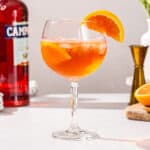 Side view of Negroni Spritz in a tall stemmed cocktail glass with orange slices as garnish. A bottle of campari, a jigger and cut orange are seen in the background.