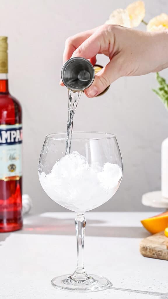 Hand pouring gin from a jigger into a cocktail glass filled with ice.