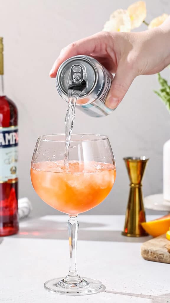 Hand pouring seltzer from a can into a red colored cocktail.