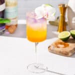 Side view of Passion Fruit Spritz cocktail on a countertop. A cut lime, Chinola liqueur and empress gin bottles are in the background, along with a jigger and purple orchid flowers. A white orchid and lime slice garnish the drink.