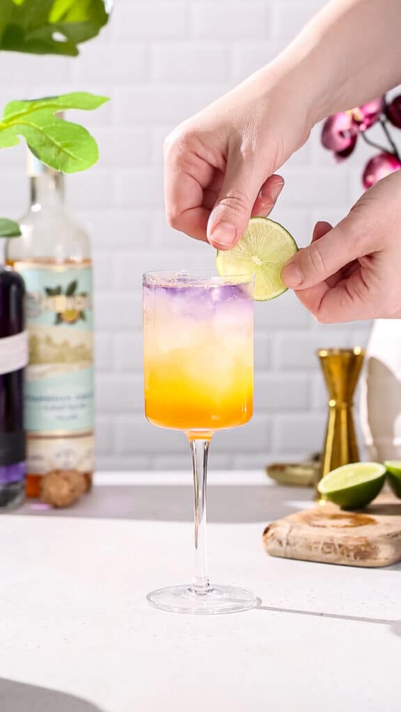 Hand adding a lime slice to a purple and orange cocktail as garnish.