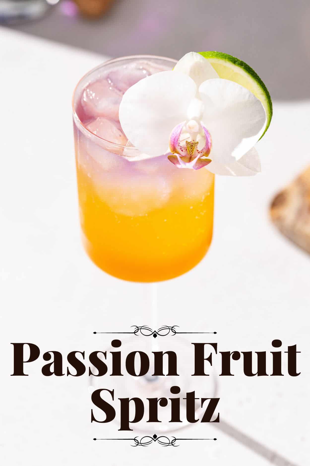 Top view of Passion Fruit Spritz cocktail, with a beautiful orchid and lime garnish. The drink is orange at the bottom with a purple color on top. Text at the bottom of the photo says “passion fruit Spritz”.
