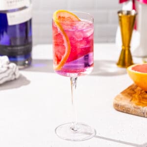 Side view of Empress Gin and tonic cocktail in a stemmed wine glass. The drink is pink and purple in color and is garnished with a pink grapefruit slice. In the background are a cut grapefruit, a jigger, and a bottle of Empress 1908 gin.