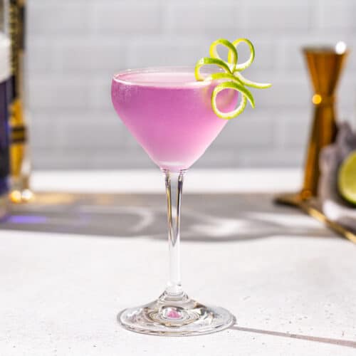 French Gimlet cocktail on a countertop in a stemmed cocktail glass with a long curly lime peel garnish. The drink is purple in color and there is a bottle of empress gin and st germain in the background along with a gold jigger and a cut lime.