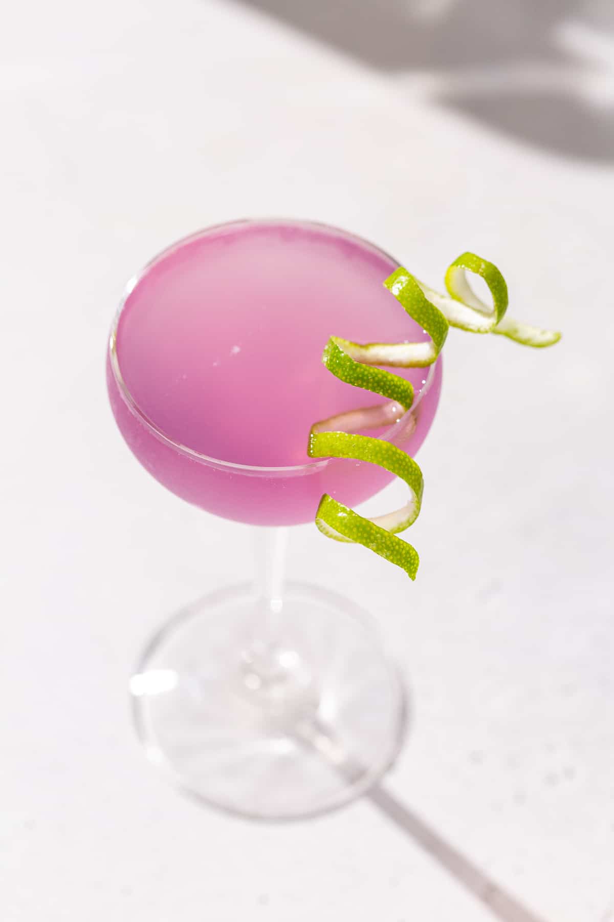 Overhead view of a French Gimlet cocktail on a countertop in a stemmed cocktail glass with a long curly lime peel garnish. The drink is purple in color.