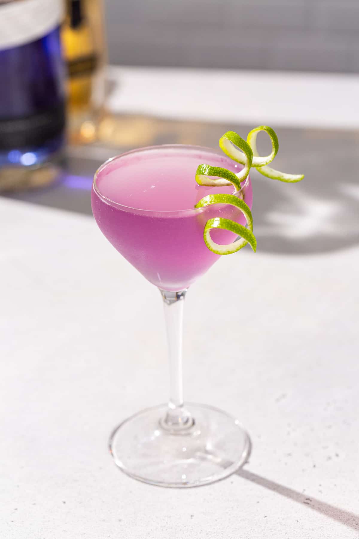 French Gimlet cocktail on a countertop in a stemmed cocktail glass with a long curly lime peel garnish. The drink is purple in color. There is a bottle of empress gin and st germain in the background.