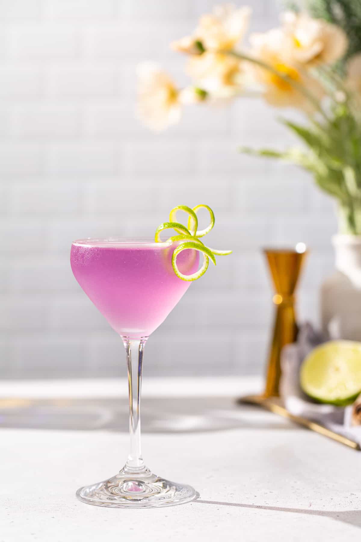 Side view of a French Gimlet cocktail on a countertop in a stemmed cocktail glass with a long curly lime peel garnish. The drink is purple in color. In the background are a gold jigger, cut lime and flowers in a vase.